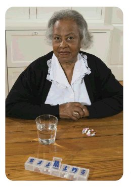 Photo of woman seated behind a glass of water, pills, and a box labeled with days of the week